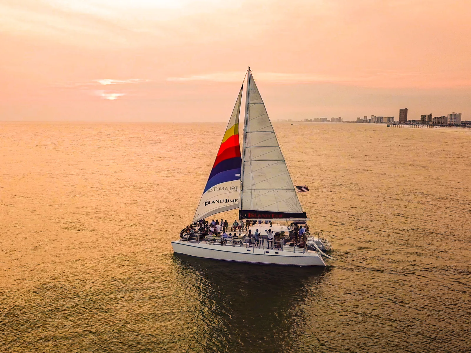 A catamaran sailing at sunset off Panama City Beach, with the developed shore in the distance.